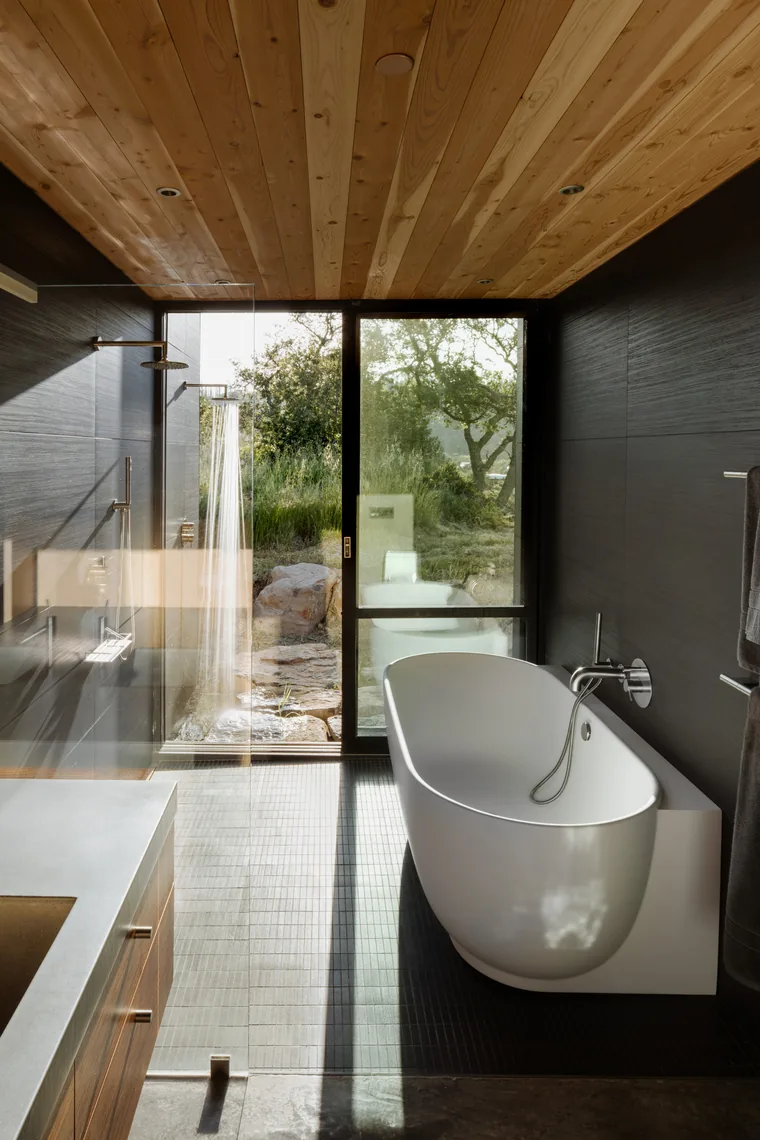 A contemporary bathroom features a freestanding tub, walk-in shower, wood ceiling slats, and a large glass door opening to an outdoor view.