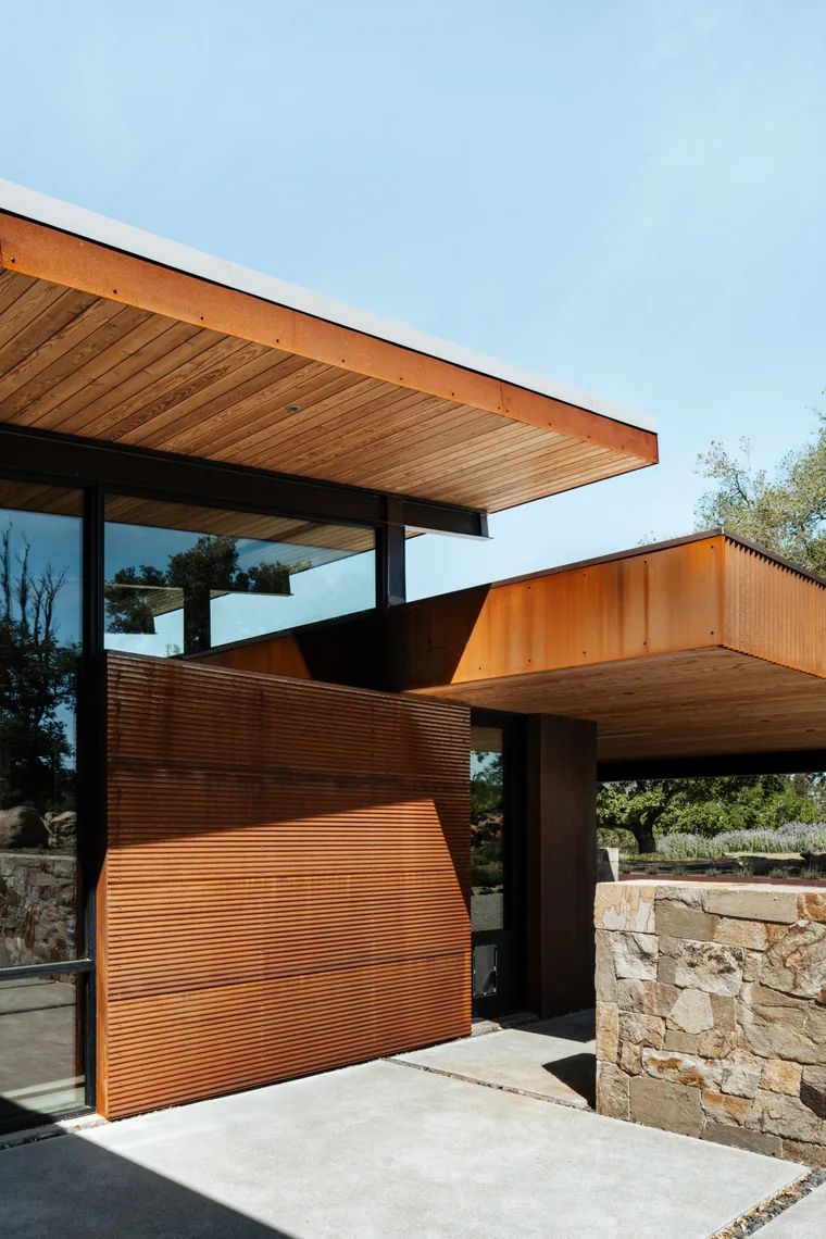 Exterior detail of HOUSE FOR A NEW BEGINNING showing wood slat siding, rusted metal cladding, and stone wall elements.