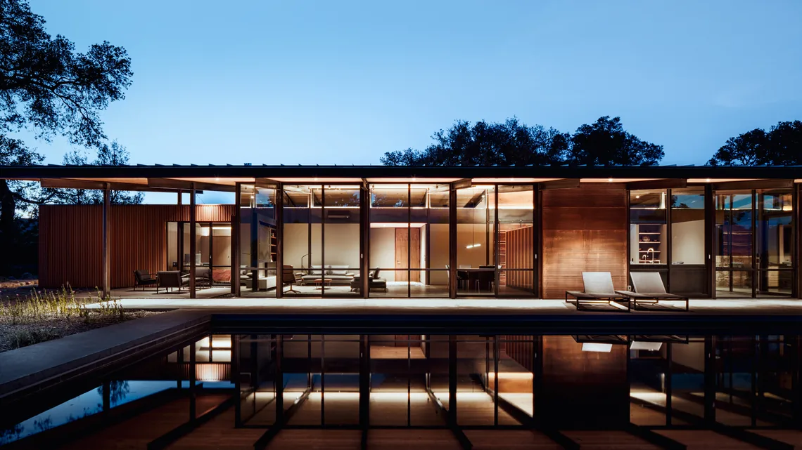 House for a New Beginning by Nielsen Schuh Architects features expansive glass walls, wood paneling, and a pool reflecting the building's warm light at dusk.