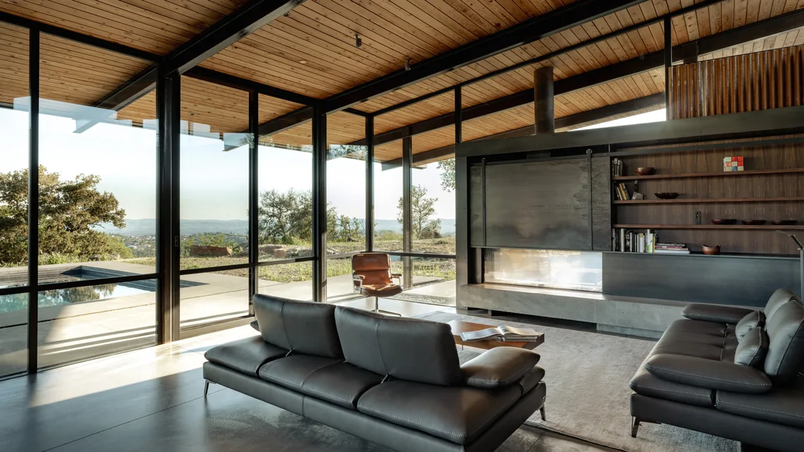 Living room with floor-to-ceiling glass walls providing pool and landscape views, featuring dark leather sofas and a wood slat ceiling.