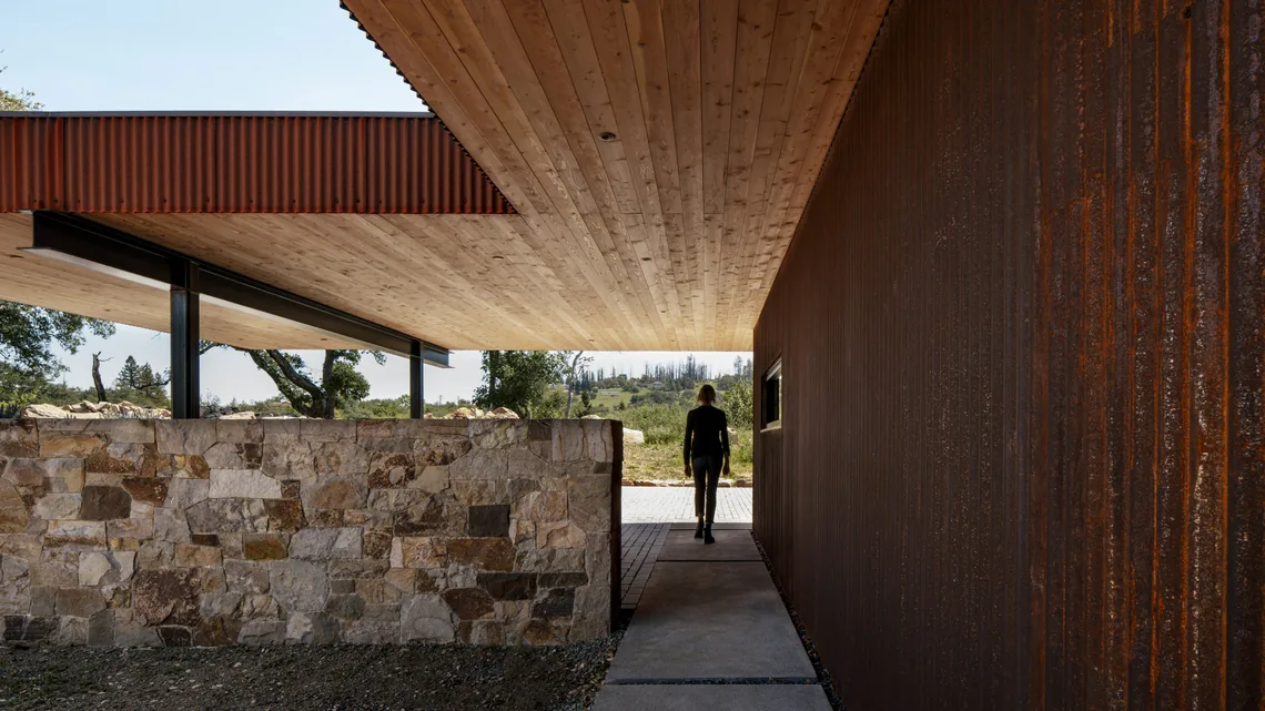 Passageway under wood ceiling with rusted steel and stone exterior walls at 'HOUSE FOR A NEW BEGINNING'.