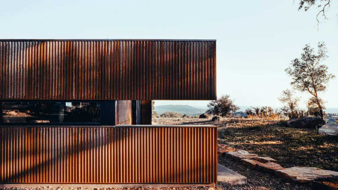 Modern home's rusted steel exterior with horizontal window strip and dry landscaped yard in sunlight.