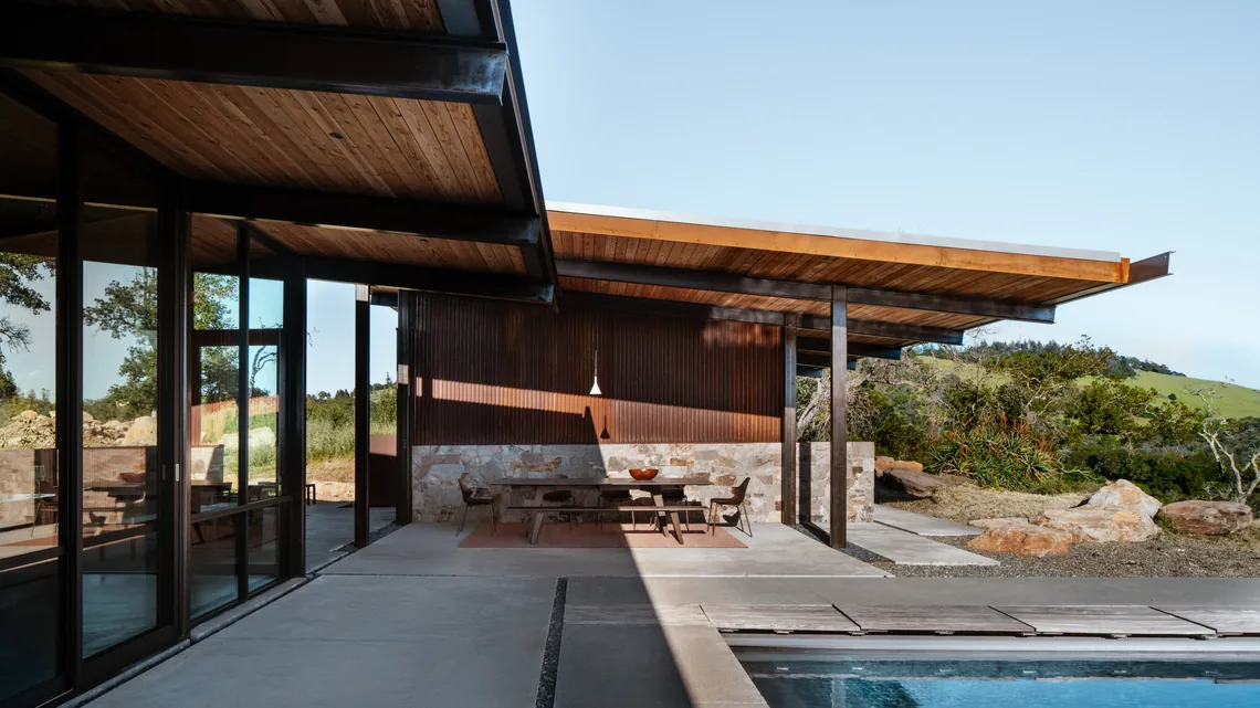 Covered patio with wood-paneled ceiling, stone accents, and seating by a pool at HOUSE FOR A NEW BEGINNING.