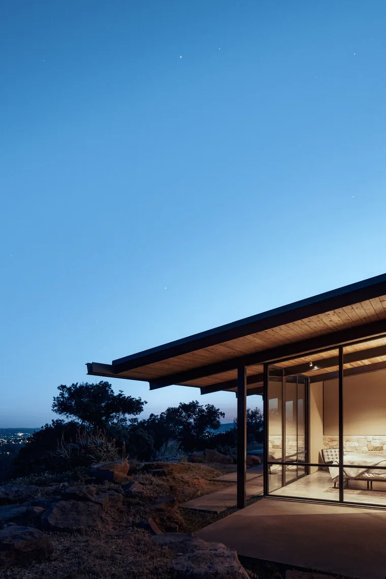 HOUSE FOR A NEW BEGINNING features metal support columns, floor-to-ceiling glazing, and a wood-paneled ceiling in a scenic hillside location.