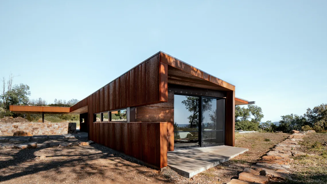 The HOUSE FOR A NEW BEGINNING features rusted steel siding and a stone half wall in a dry, natural landscape.
