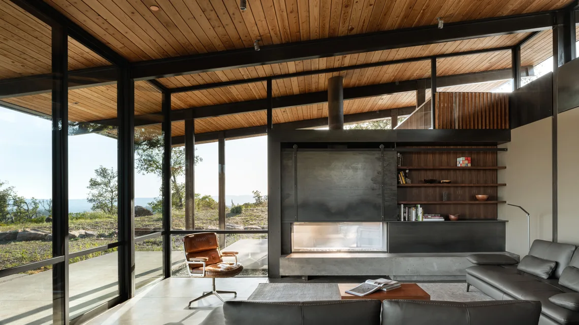Interior living space featuring large glass walls, a leather lounge chair, and a built-in modern fireplace under a wood slat ceiling.
