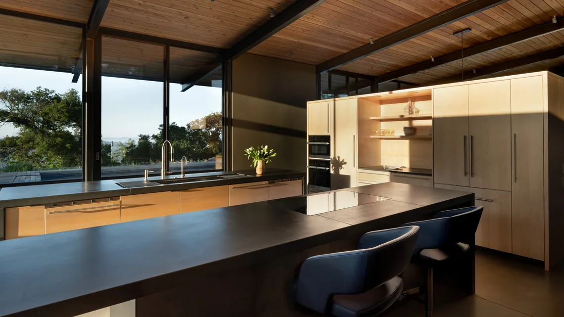 Modern kitchen with wood cabinetry, large island, and floor-to-ceiling windows overlooking outdoor scenery.