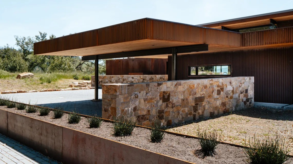 Modern house exterior with rusted steel siding, stone retaining walls, and paved driveway features in a dry grass setting.