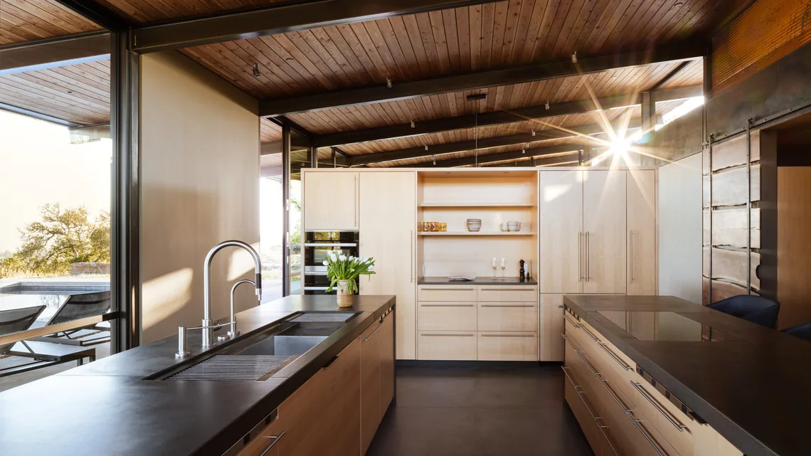Kitchen with wood cabinets and black countertops featuring a double sink and induction cooktop, opening to an outdoor area with pool loungers.