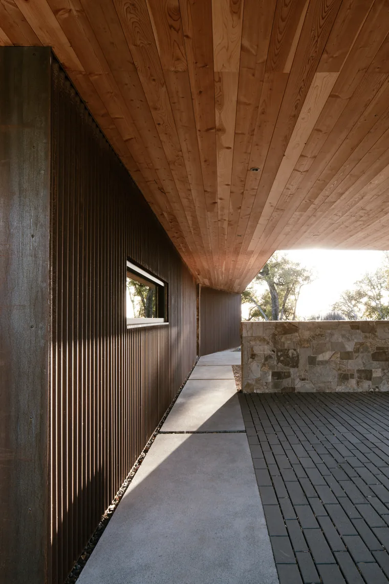 Covered walkway with dark vertical wood siding, wood ceiling, concrete path, and stone wall enclosure.