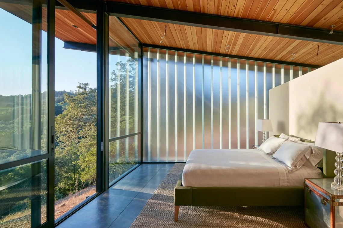 Modern bedroom features floor-to-ceiling glazing, sloping wood ceiling, and frosted glass panel wall for privacy.
