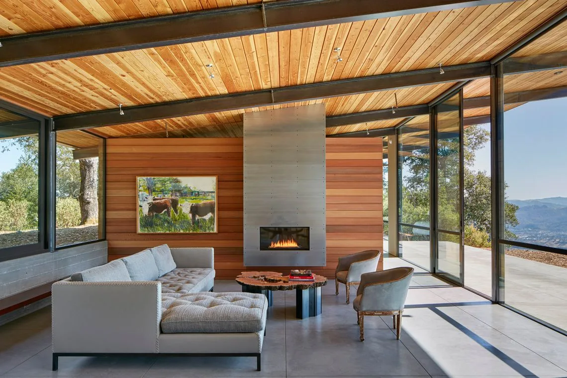Living room of Split Rock Springs Ranch features floor-to-ceiling glass walls and a sleek metal fireplace against a wood-paneled wall.