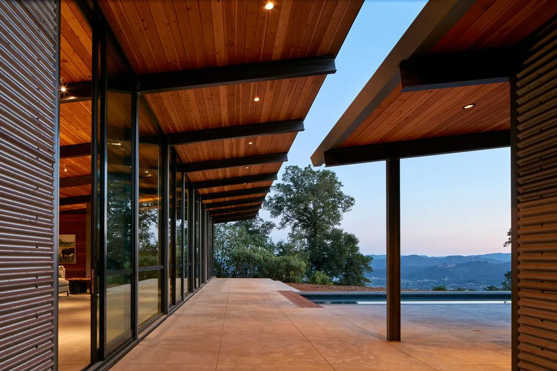 The home features a covered patio with wood-paneled ceilings, black steel beams, and floor-to-ceiling glass walls overlooking a pool.