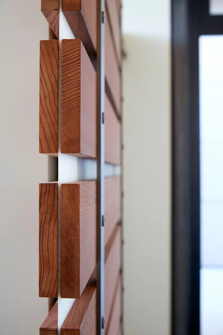 Detail of wooden blocks mounted in a grid on a white panel wall adjacent to a black metal-framed window.