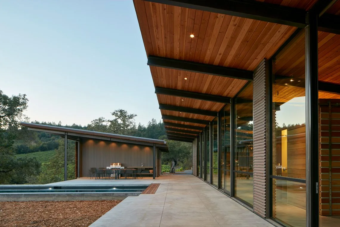 Covered patio with a wood-paneled ceiling, floor-to-ceiling glazing, and an outdoor dining area under a sloping roof.