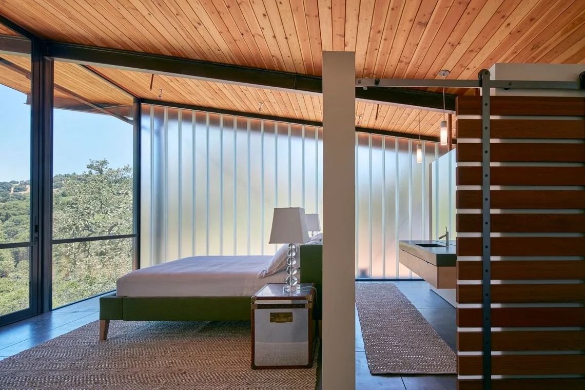 Bedroom with floor-to-ceiling frosted glass wall, wooden ceiling, and sliding glass door opening to a wooded landscape.