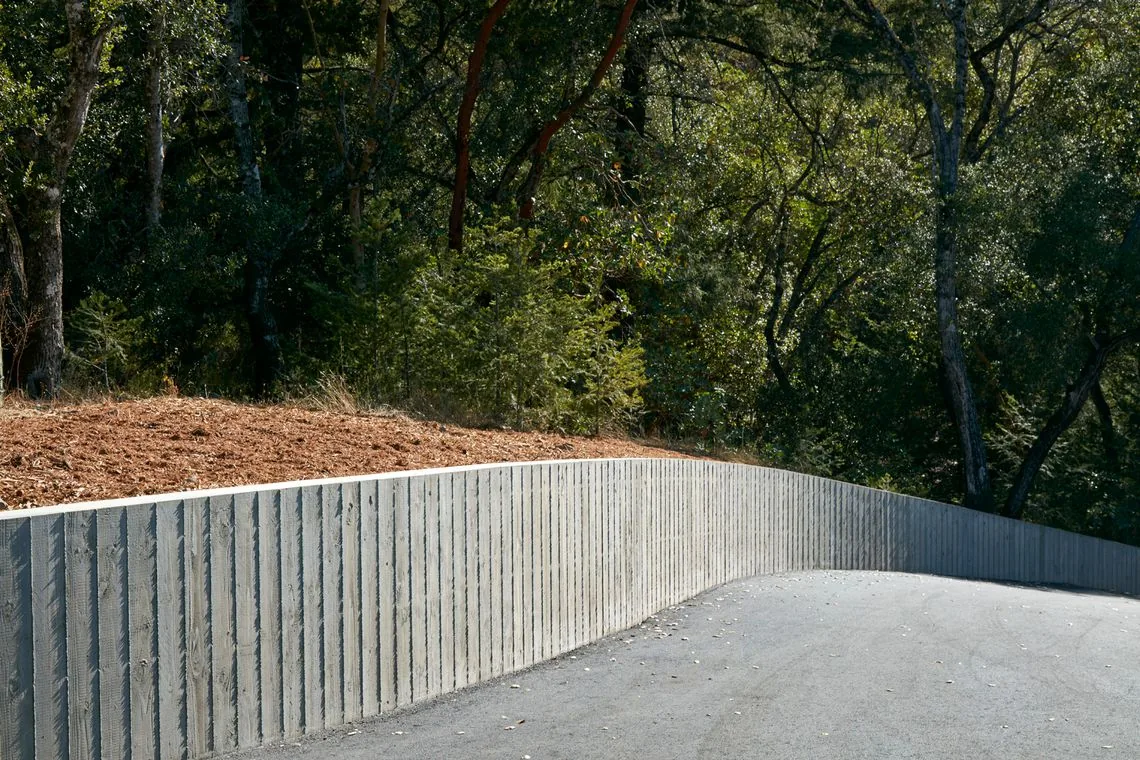 Board-formed concrete retaining wall curves along the edge of an asphalt driveway beside wooded terrain.