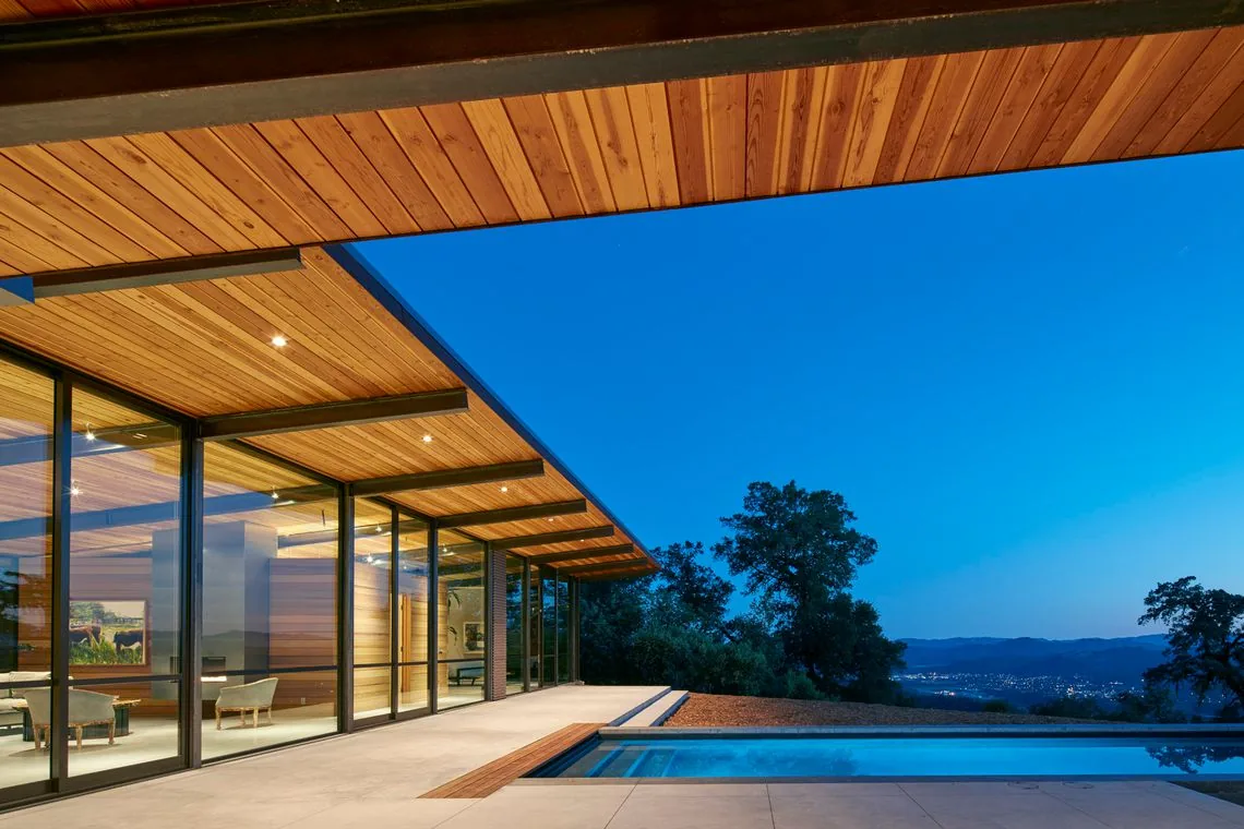 Split Rock Springs Ranch features a modern home with expansive glass walls, wooden ceiling overhang, and pool overlooking wooded hills.