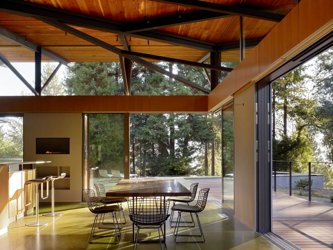 Open dining and kitchen area with wire chairs, wood table, and sliding glass doors opening to a raised deck with forest views.