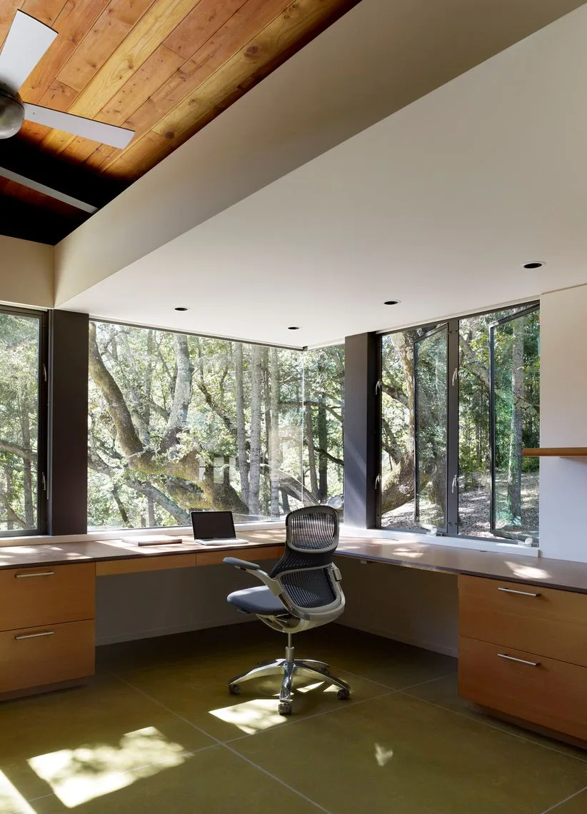 Home office features a wood L-shaped desk with ample storage and large corner windows providing forest views.