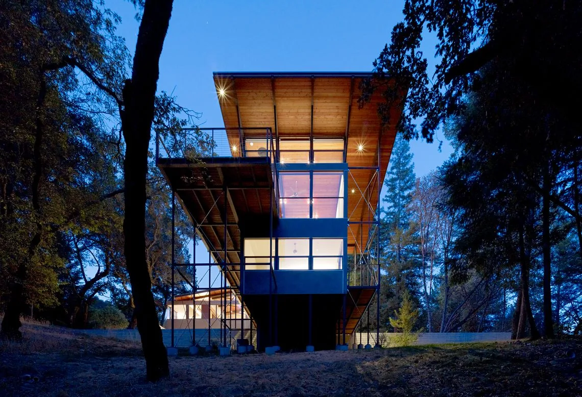 Sonoma Mountain House features an overhanging roof with metal bracing, set on a raised foundation in a forest landscape.