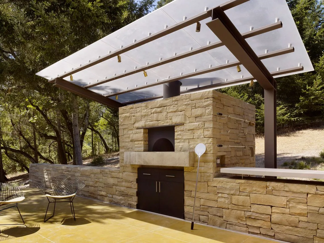 Outdoor kitchen with stone pizza oven and metal pergola in a forested area of Sonoma Mountain House project.