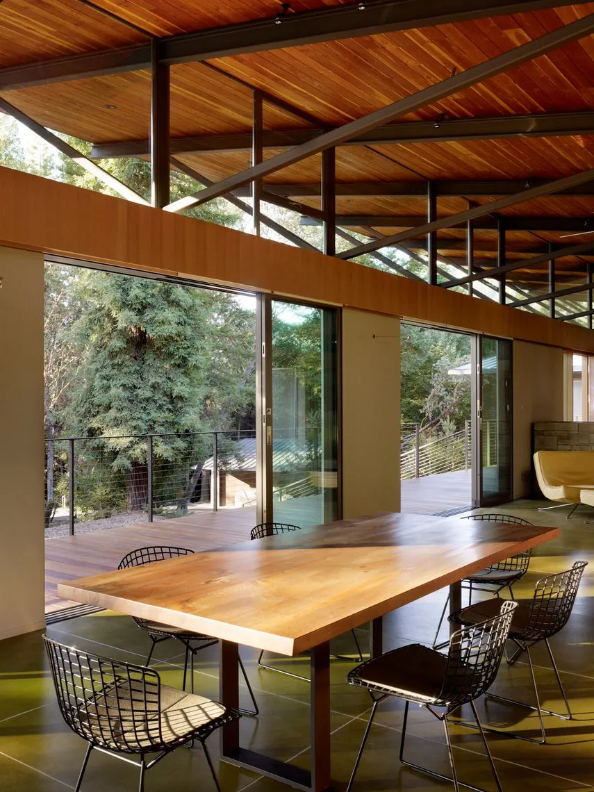Dining room features a wood table with metal wire chairs and floor-to-ceiling sliding glass doors overlooking a wooded exterior deck.