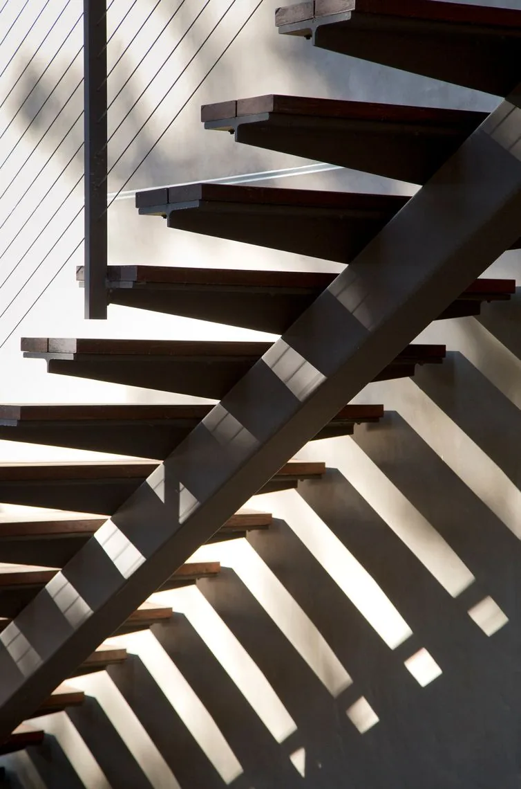 Detail of a floating staircase with dark wood treads and metal stringer, showing shadow patterns on the adjacent wall.