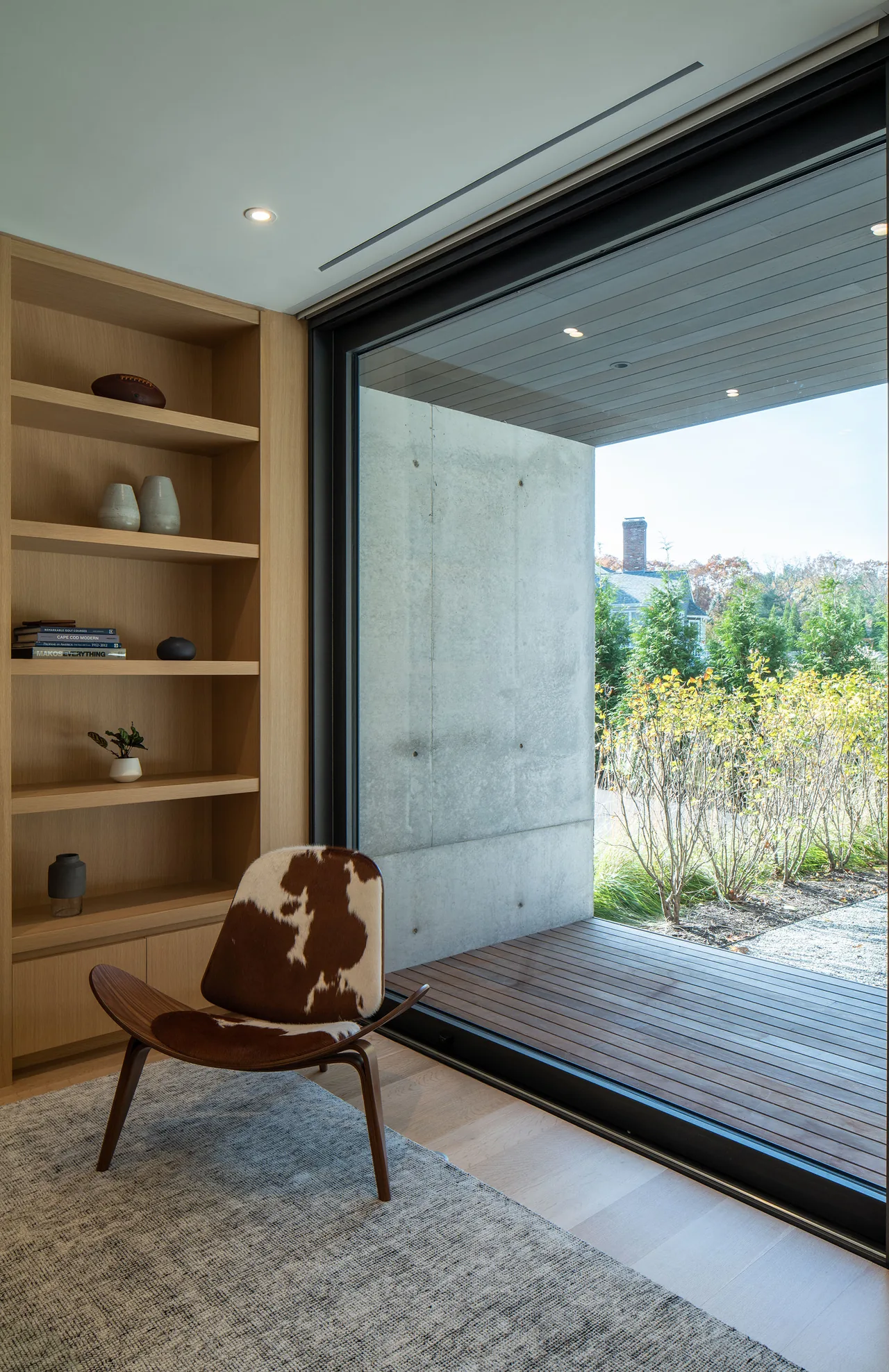 Room corner features light wood built-in shelves and a cowhide chair beside floor-to-ceiling sliding glass door opening to a wooden deck.