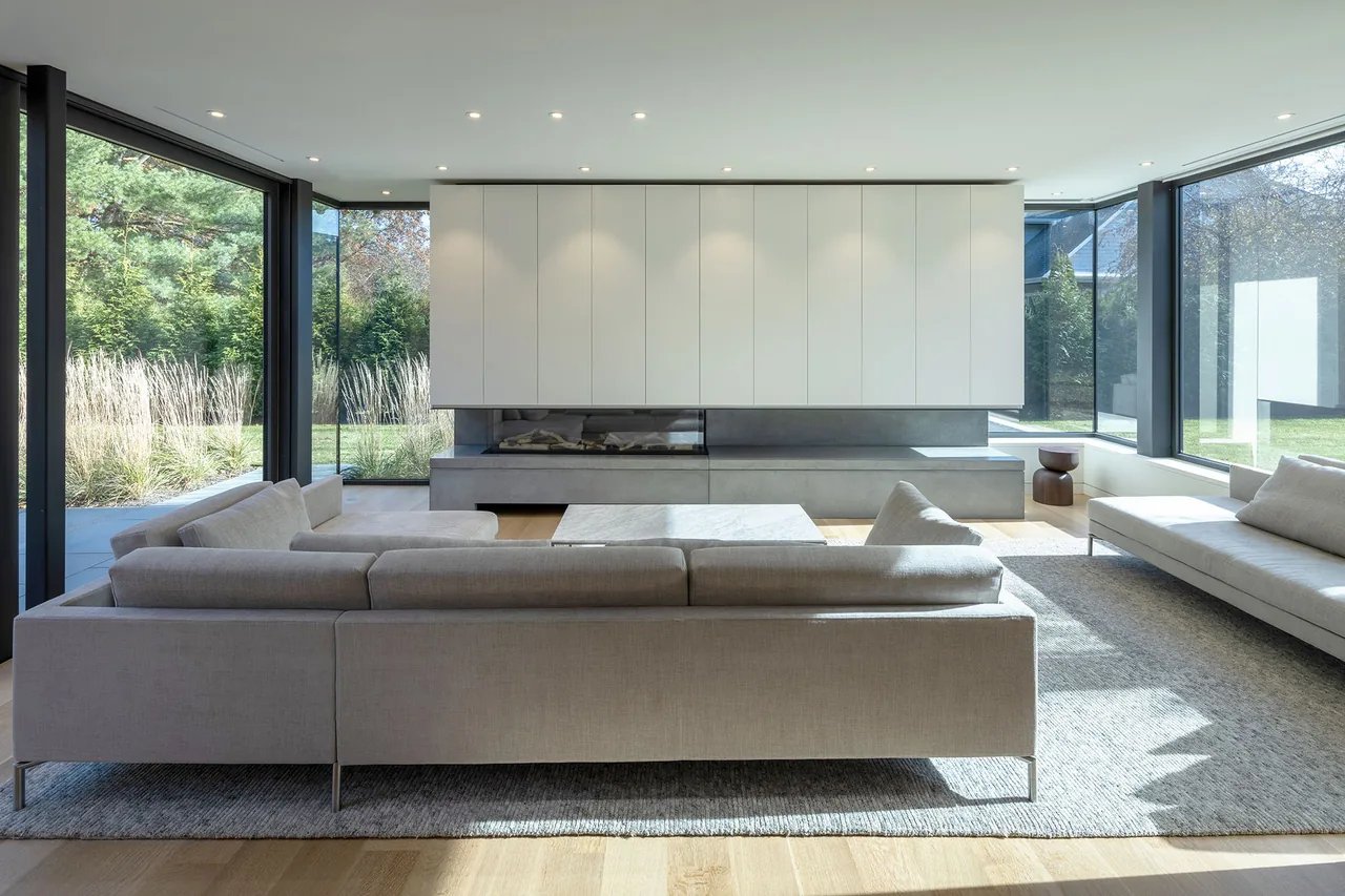 Living room with beige sectionals, floor-to-ceiling glass walls, and a minimalist built-in fireplace feature.