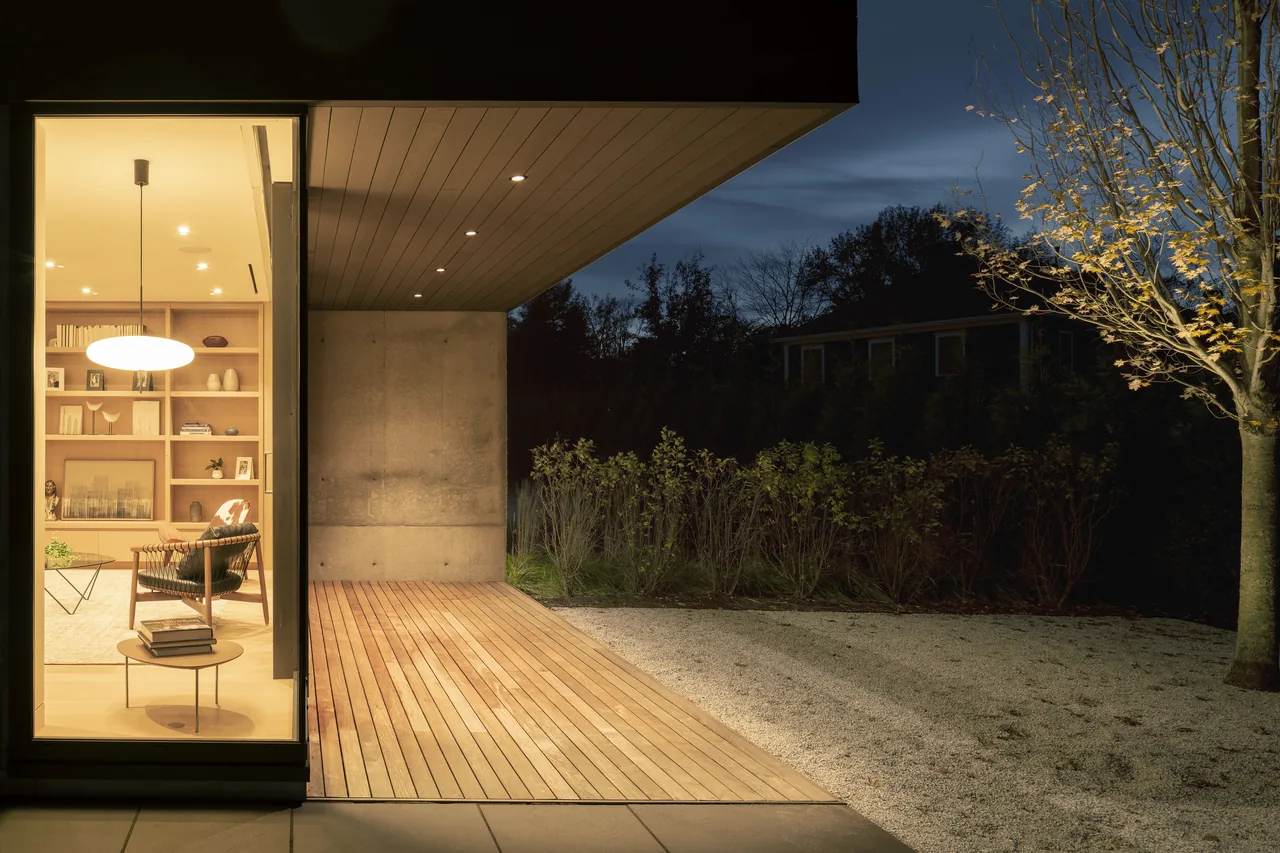 Modern house exterior with wood decking, a concrete corner wall, and an illuminated interior living room with built-in wooden shelving.