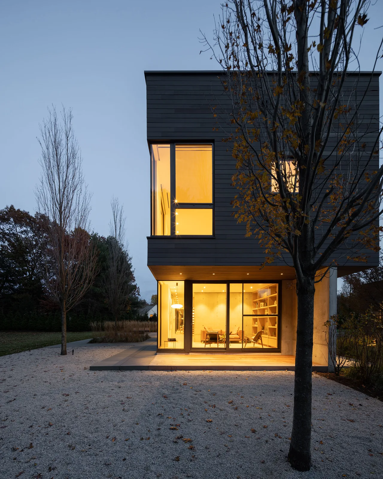 NEEDHAM HOUSE features a modern exterior with floor-to-ceiling glazing illuminating the interior at dusk.