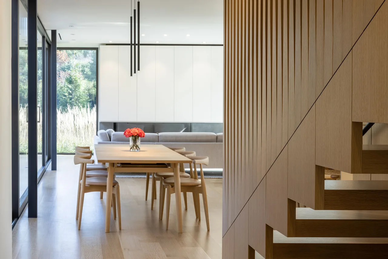 Modern dining area with light wood furniture, floor-to-ceiling glazing, and a visible wooden staircase detail.