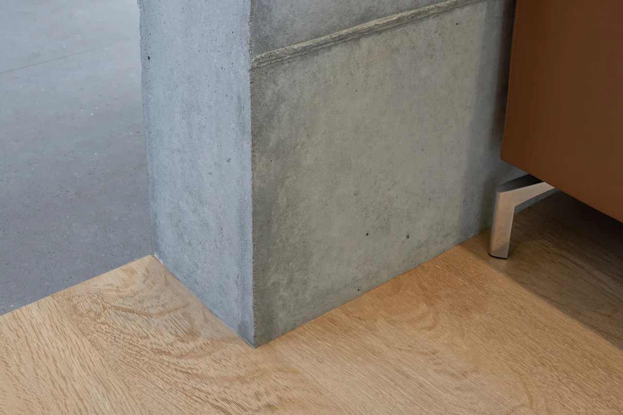 Detail showing the meeting point of textured concrete wall and light wood floor with a sofa leg visible.