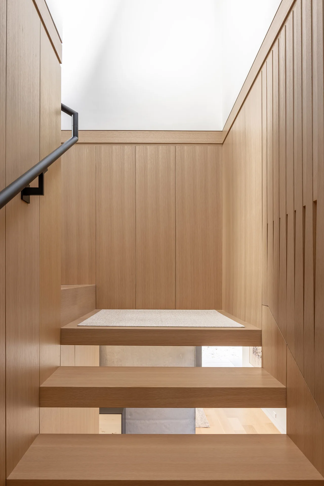 Light wood floating staircase next to matching wood paneled walls with a black metal handrail.
