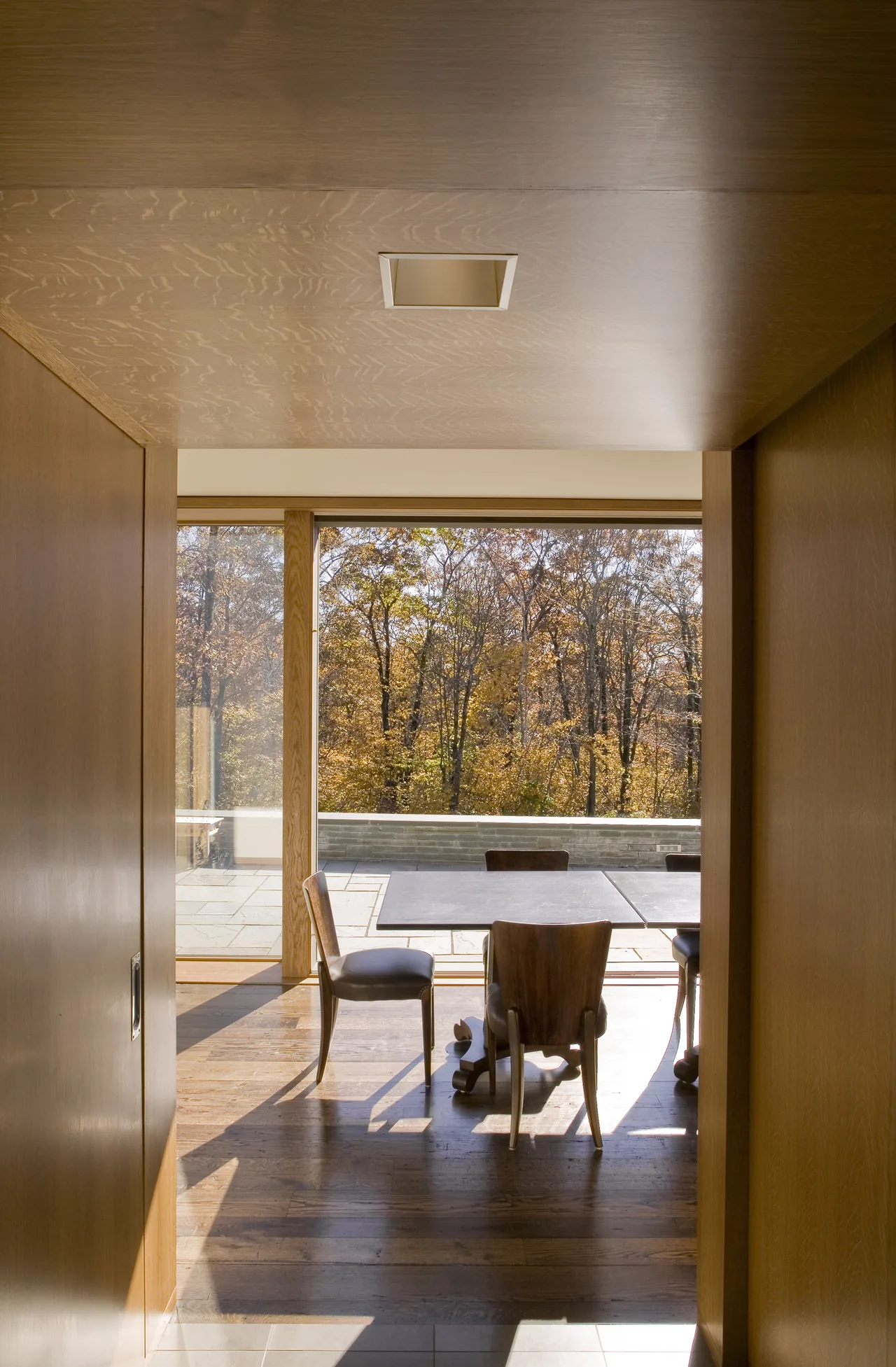 Wood-paneled hallway leads to a dining area with wooden chairs and large glass windows overlooking fall trees.