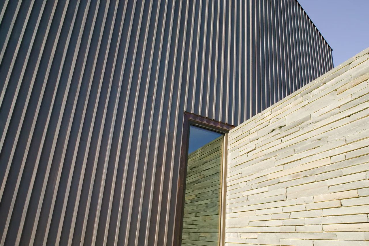 The Blue Rock House exterior features vertical metal siding juxtaposed with a stone wall and a rectangular window.