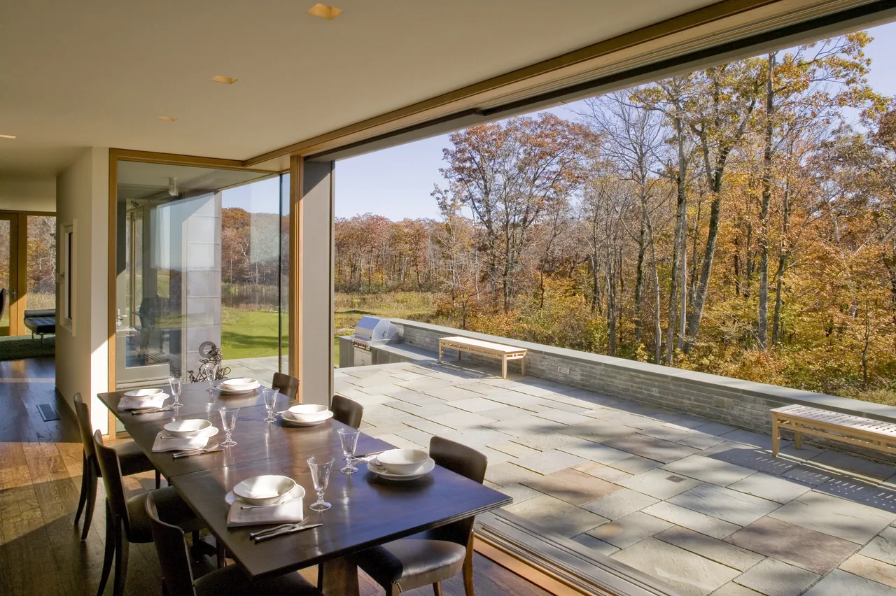 The dining room opens to a large stone tile patio with benches and a built-in grill, surrounded by trees in autumn.