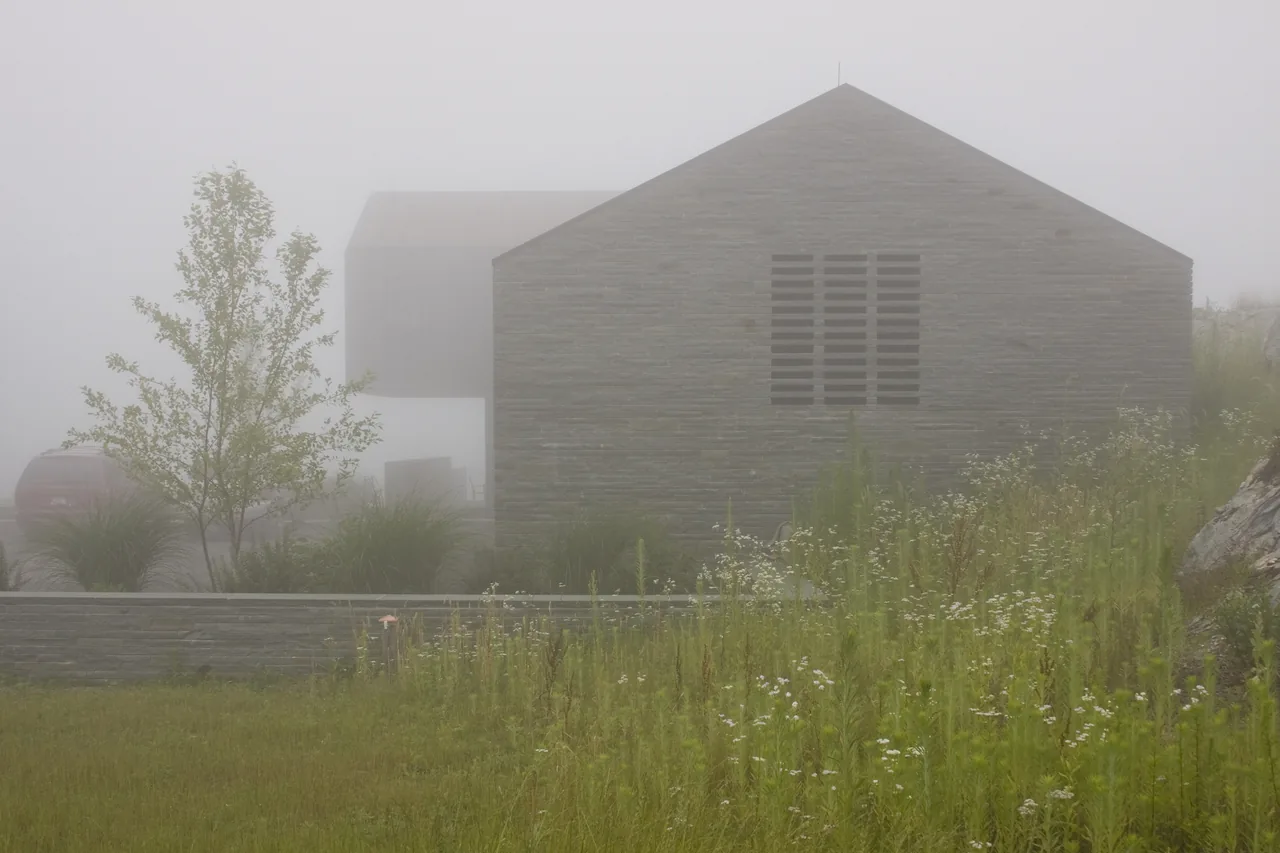 Exterior view of a stone building with a gable roof and a stone retaining wall surrounded by natural landscaping in foggy conditions.