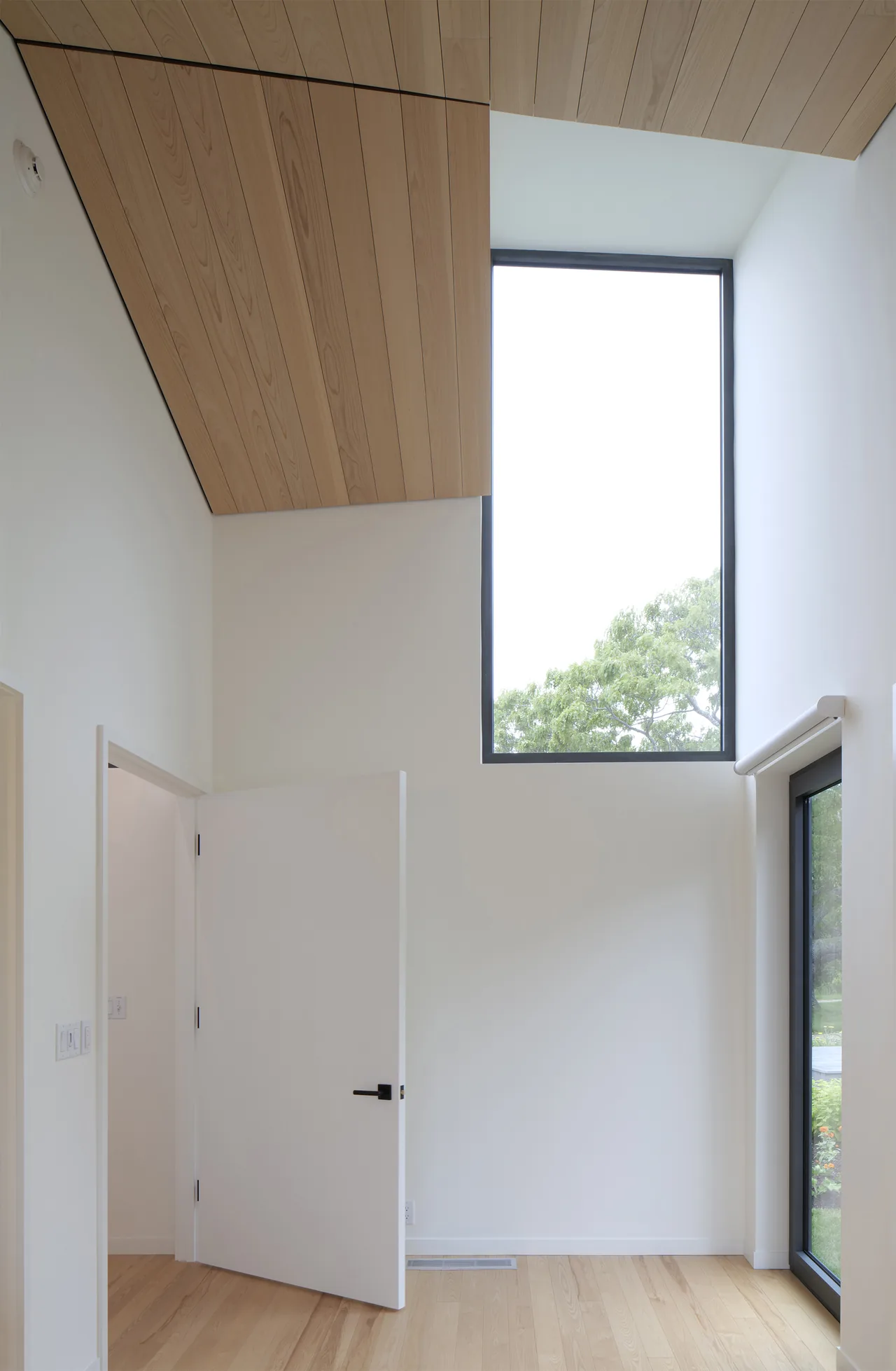 Interior corner shows wood-paneled ceiling, black window frame, and white door with black handle opening into another white-walled space.