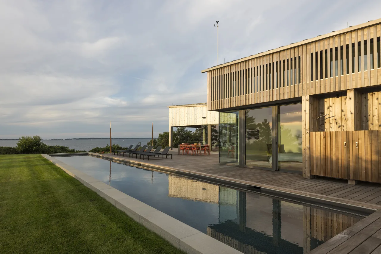 The TWO SHEDS HOUSE features a rectangular pool next to a wood exterior with a covered porch overlooking a waterfront.