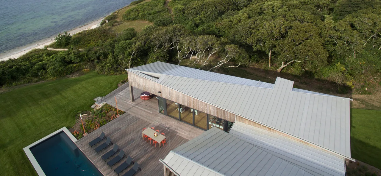 The TWO SHEDS HOUSE features standing seam metal roofing, a wood deck, and an outdoor pool next to a waterfront site.