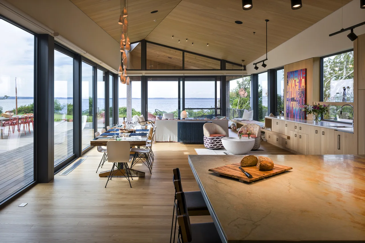 The TWO SHEDS HOUSE features an open living and kitchen area with wood ceiling panels and expansive glass walls overlooking the waterfront.