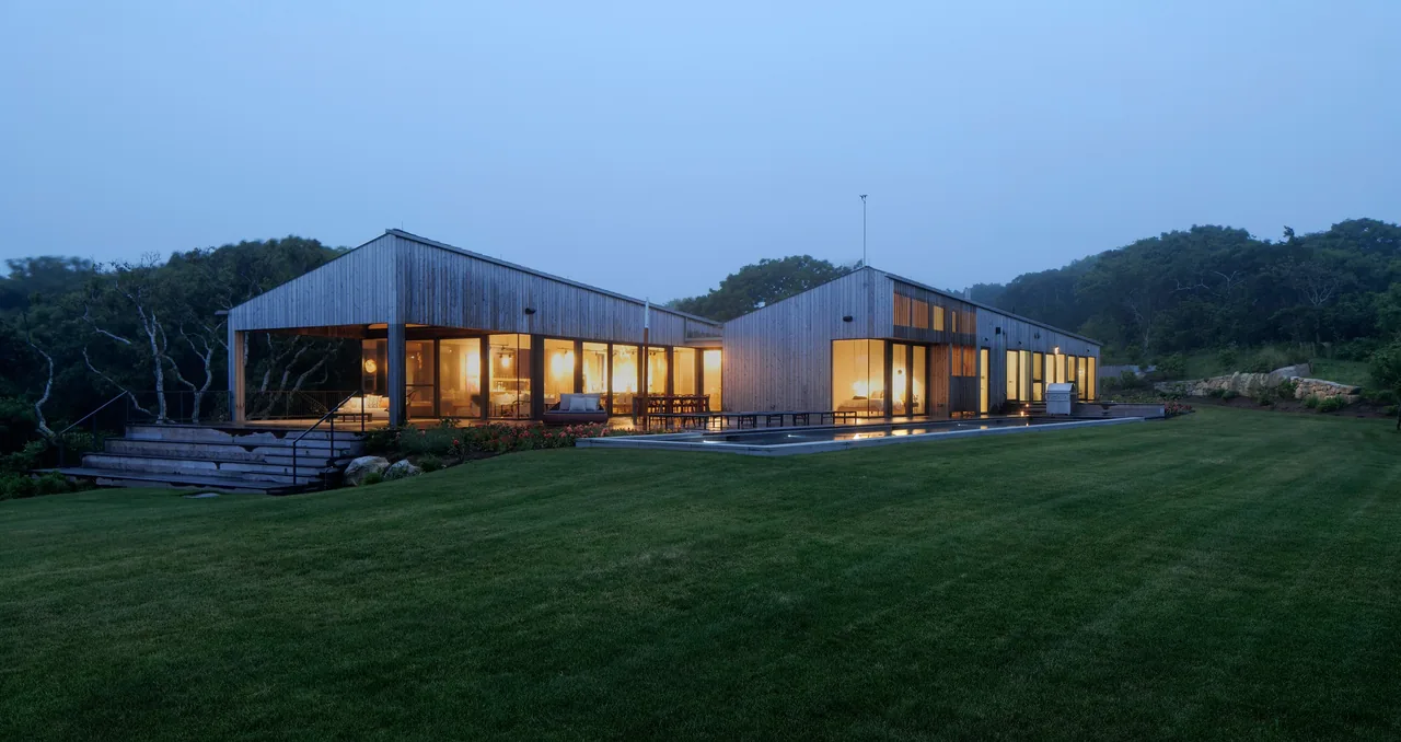 The TWO SHEDS HOUSE features two connected shed-style structures with extensive glazing and wood siding, illuminated at dusk.