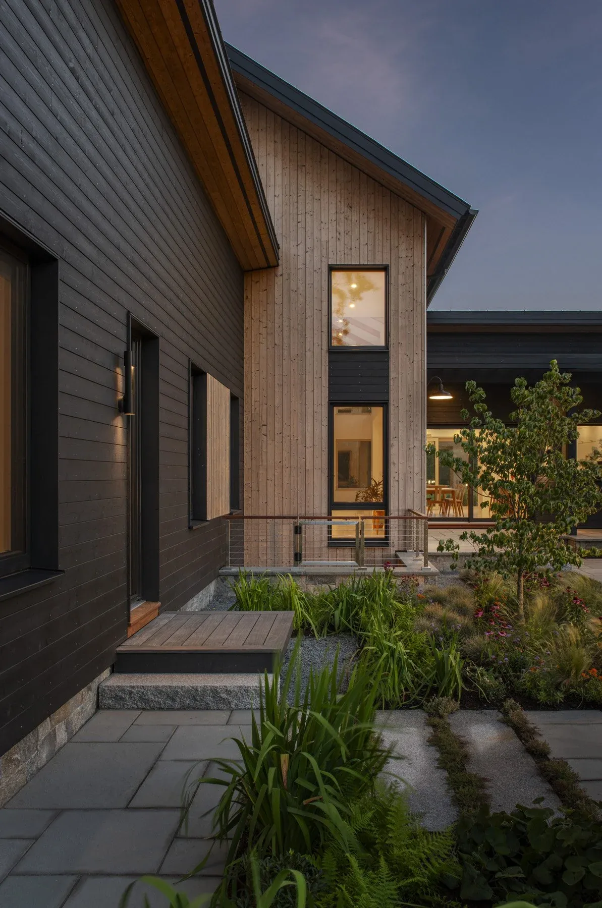 House exterior combining black horizontal wood siding with natural vertical wood siding and stone accents, featuring a landscaped garden with a small tree.