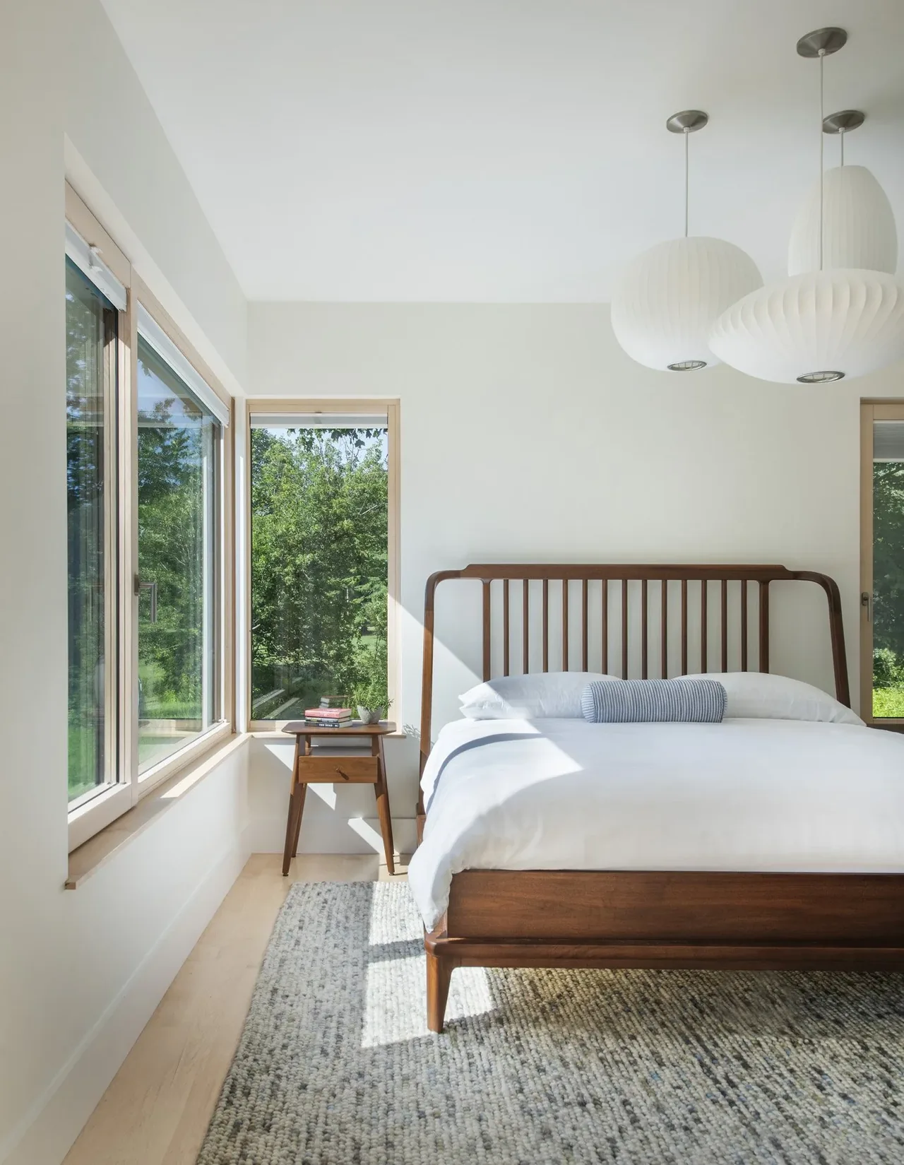 A bright bedroom featuring a wooden bed with a slatted headboard and large corner windows overlooking greenery.