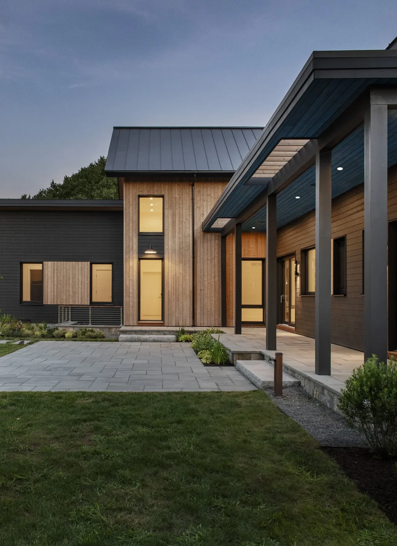 Stow Passive House features wood siding, black window frames, and a covered patio with stone pavers in Boston.