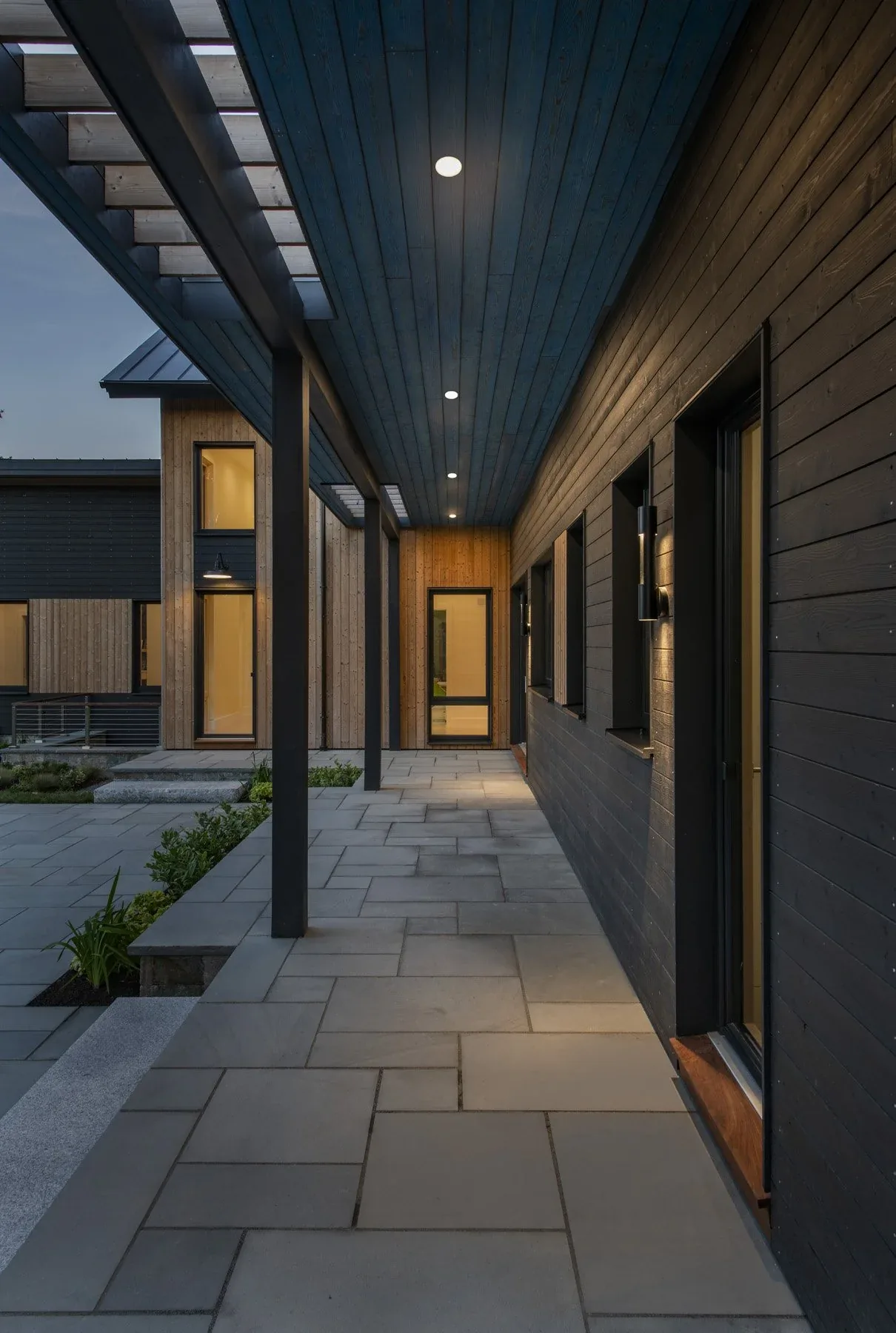 Covered exterior walkway of the Stow Passive House features black wood siding, stone tile floor, and recessed ceiling lights.