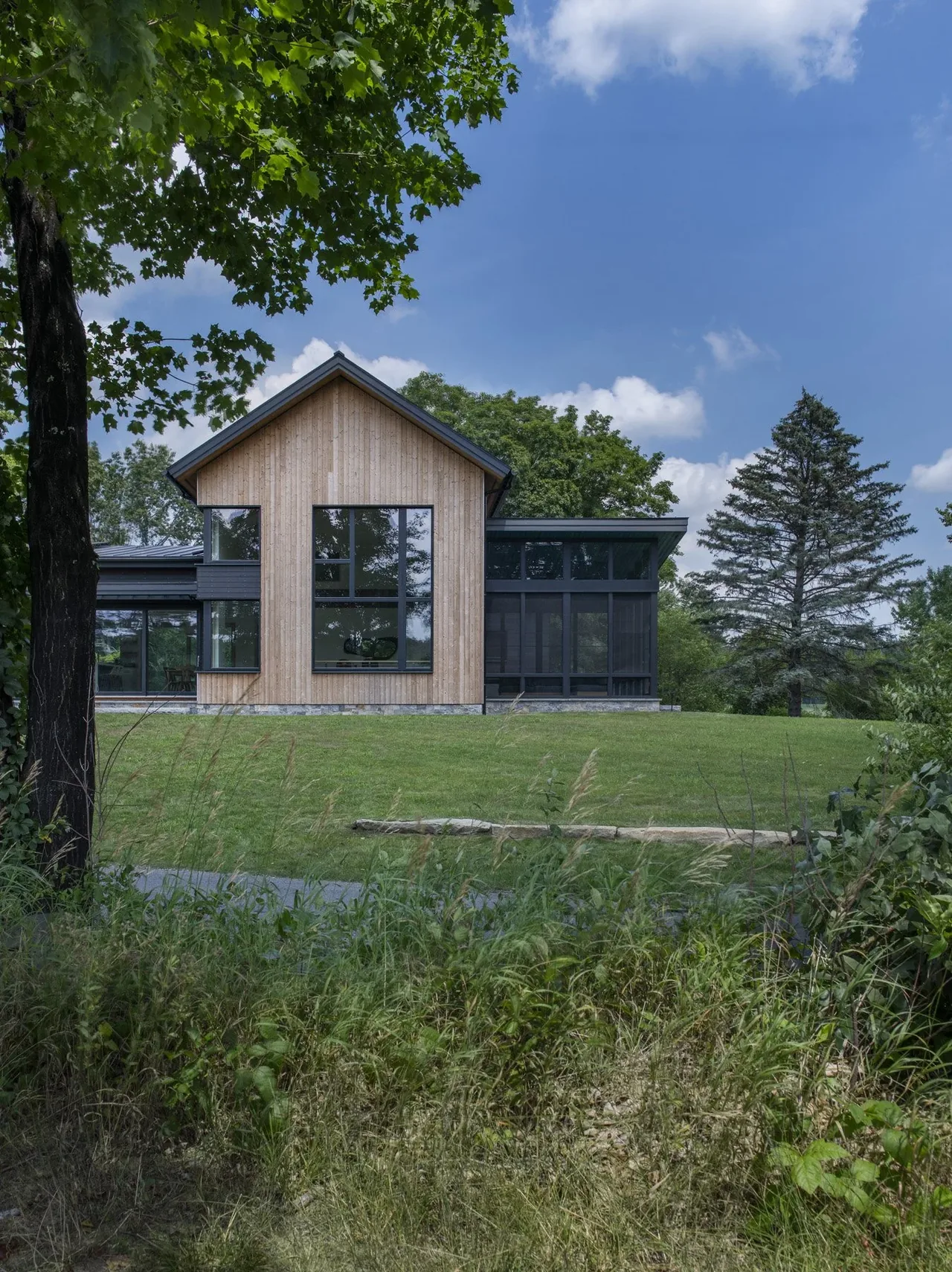 Modern wood-clad house with large black-framed windows and gable roof surrounded by greenery.