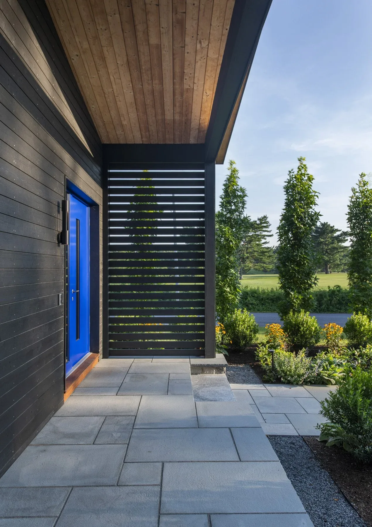 The Stow Passive House entryway features vertical black siding, a vivid blue door, and a wood-planked ceiling over a stone-paved floor.